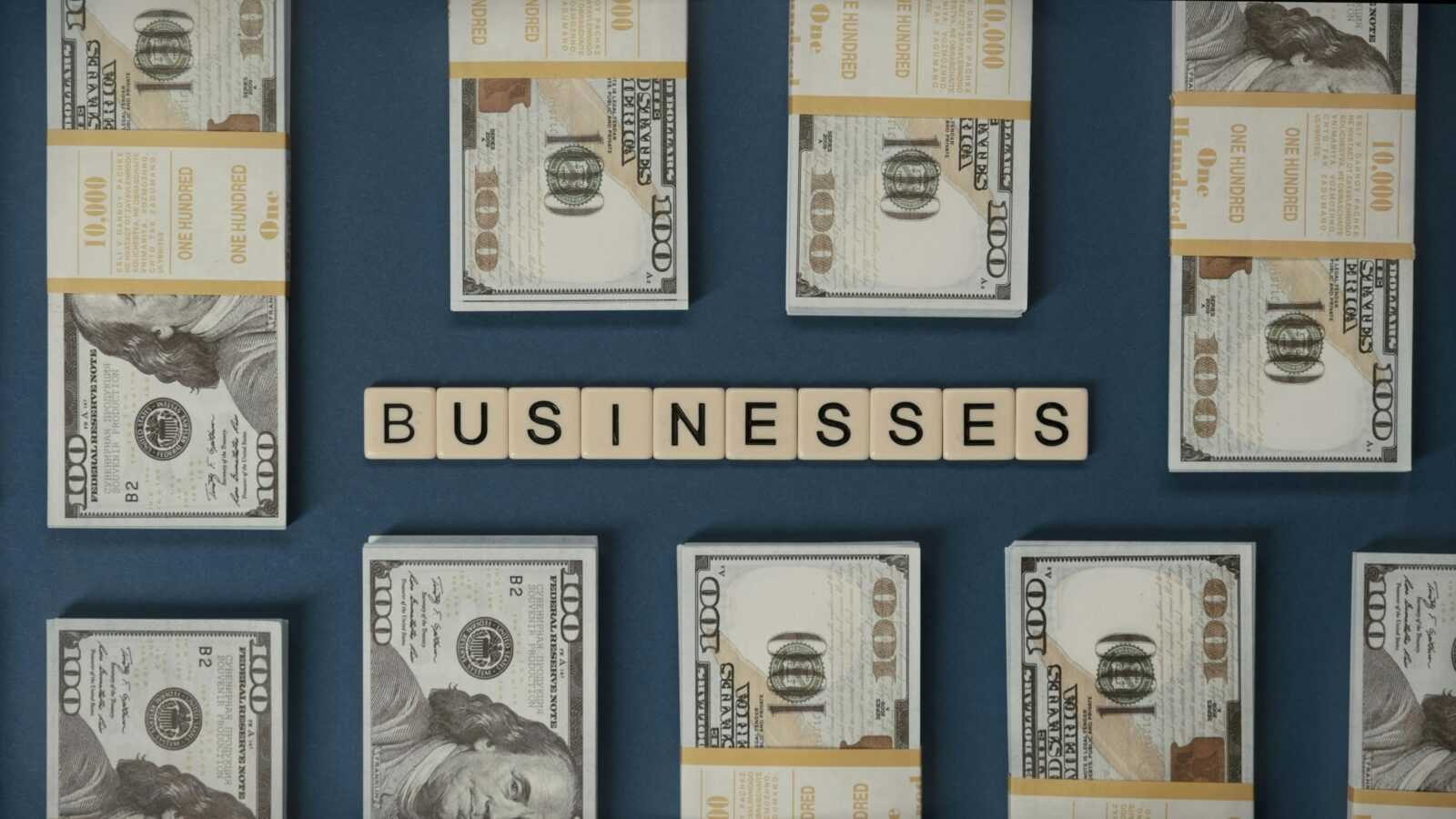 Bundles of US hundred-dollar bills and Scrabble tiles spelling 'businesses' laid out creatively.