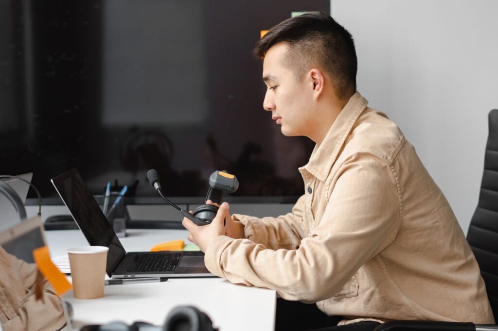 Professional Asian man working on laptop in a modern office setting, using a headset.